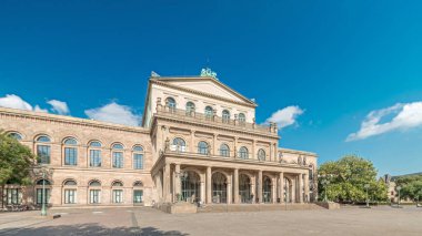 Hanover Devlet Operası 'nın (Staatsoper Hannover) ön görüntüsü. Şehir meydanında canlı mavi bulutlu bir zaman dilimi altında yürüyen insanların olduğu klasik tarzda bir tiyatro. Almanya