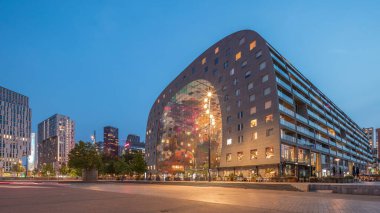 Rotterdamse Markthal 'ın dış görünüşünün geceden güne geçişi. Dükkanları ve restoranları olan ikonik bir bina. Meydanda gezinen insanlar. Gökdelenler ve arka planda canlı bir şehir manzarası