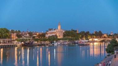 İspanya, Sevilla 'daki Torre del Oro gözlem kulesi. Günbegün geçiş zamanı, şehrin ışıkları su ve deniz manzarasını yansıtıyor. Guadalquivir Nehri boyunca yüzen bir tekne.