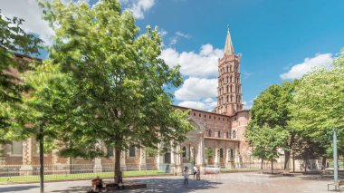 Fransa 'nın Toulouse kentindeki Saint Sernin Bazilikası' nın kaldırımlı parvisleri. Zaman atlaması. Avrupa 'nın en büyük Roma kilisesi tuğla çan kulesi, kemerli kapılar, yeşil ağaçlar ve mavi bulutlu gökyüzü.