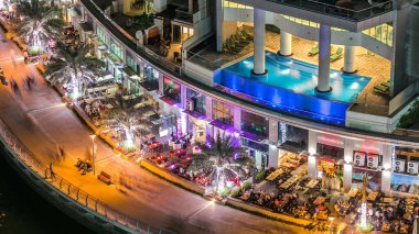 Dubai Marina 'nın gece gezintisi sırasında Dubai, BAE' de yüzen yatları ve tekneleri olan restoran ve kanallarla hava manzaralı. Yanıp sönen ışıkları ve yürüme alanı olan aydınlık modern kuleler