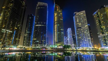Rıhtımdaki ışıklandırılmış gökdelenlerin gece görüş açısını hızlandırın. Dubai, BAE 'deki Jumeirah Gölü Kuleleri' ndeki konut binaları. Rıhtımdan suya yansıyan görüntüyü göster