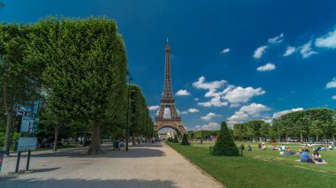 Paris 'teki Champs de Mars' taki Eyfel Kulesi. Zaman atlaması, Fransa. Yazın mavi bulutlu gökyüzü Ağaçlı yeşil çimenler ve etrafta yürüyen insanlar.