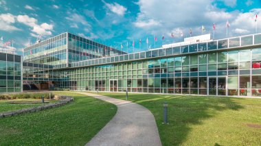 İş bölgesindeki modern düşük katlı cam ofis binası ve iç avlu zaman aralığında hızlandırılmış yol olan yeşil çimenler. Çatıda pek çok farklı ülkenin bayrakları