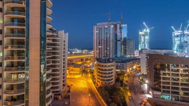 Dubai Marina 'da aydınlık gökdelenler ve gece gündüz gezinti güvertesi geçiş zamanı. Yol trafiği olan modern kuleler ve tekneler. Birleşik Arap Emirlikleri