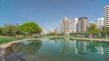 Abu Dabi 'nin sahil şeridi boyunca uzanan Corniche Bulvarı plajı parkında arka planda gökdelenler var. Lake Park 'tan yansıtmalı görüntü. Bir yanda ağaçlar