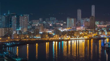 Ajman 'ın şehir manzarası gece vakti çatıdan göldeki suyun yansımasıyla görüntüleniyor. Birleşik Arap Emirlikleri 'nin rıhtımındaki aydınlık kuleler..