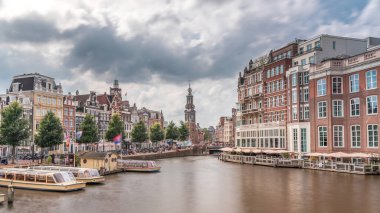 Amstel Nehri, rıhtım ve Munttoren kulesinin yer aldığı Amsterdam şehir merkezinin panoramik zaman atlaması. Tekneler bulutlu gökyüzünün altında ileri geri yüzerler. Köprüden manzaralı şehir manzarası. Hollanda