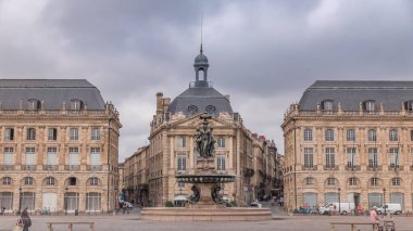 Place de la Bourse zaman atlaması Fransa 'nın Bordeaux şehrinde, Bulutlu mavi gökyüzünün altında klasik mimari ile çevrili Üç Zarafet Çeşmesi ile birlikte. İnsanlar ana meydanda bir tramvay durağında