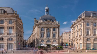Place de la Bourse, Fransa 'nın Bordeaux şehrinde, bulutlu mavi gökyüzü altında klasik mimari ile çevrili restoranları olan bir yer. Meydanda ve ana caddelerde yürüyen insanlar