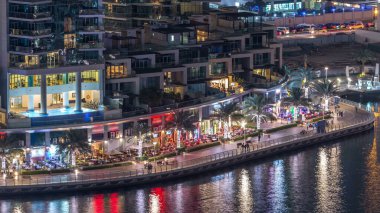 Lüks Dubai Marina kanalı kıyısı, oradan geçen tekneler ve restoranlarla dolu bir gezinti yeri, yukarıdan aydınlık gökdelenleri olan bir manzara. Dubai, Birleşik Arap Emirlikleri