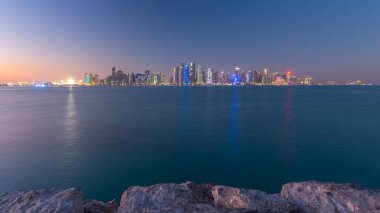 Doha şehir merkezi ufuk çizgisi günden geceye geçiş zamanı, Katar, Orta Doğu. Batı Körfezi 'ndeki aydınlık gökdelenler Körfez suyuna yansıyor.