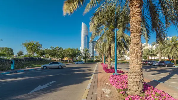 Abu Dabi 'nin sahil şeridi boyunca uzanan Corniche Bulvarı plajı parkında arka planda gökdelenler var. Aile parkından görüntü. Palmiyeler ve çiçekler yan yana. Güneşli bir günde mavi gökyüzü