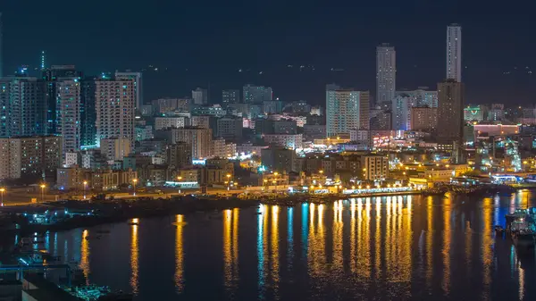 Ajman 'ın şehir manzarası gece vakti çatıdan göldeki suyun yansımasıyla görüntüleniyor. Birleşik Arap Emirlikleri 'nin rıhtımındaki aydınlık kuleler..