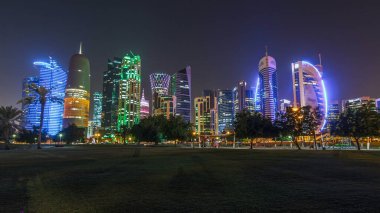Geceleri Doha 'nın ufuk çizgisi. Yıldızlı gökyüzü Park Timelapse hiper hızından görünüyor, Katar. Ön planda ağaçlar. Aydınlatılmış gökdelenler ve kuleler