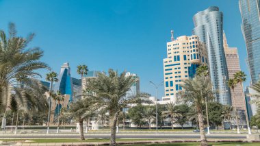 Doha 'nın ufuk çizgisi Park Timelapse, Katar' dan görüldü. Ön planda ağaçlar ve palmiyeler. Arka planda modern gökdelenler ve kuleler. Yolda trafik vardı.