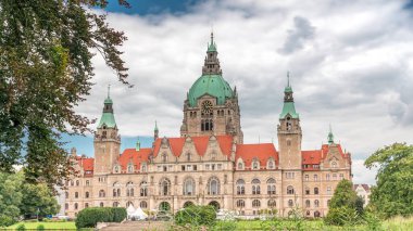 Hanover 'in Yeni Belediye Binası' nın hiperlapesi. Bulutlu bir gökyüzünün altında park ve göl ile çevrili tarihi bir kale gibi bir yapı. Su ve yeşillik üzerindeki yansımalar bu ikonik şehir manzarası zaman çizelgesini oluşturuyor. Almanya