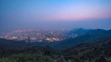 Hong Kong şehri Kowloon Tepesi 'nin tepesinde gündüz gece aydınlık gökdelenlerle aydınlatılmış şehir manzaralı bir geçiş zamanı olarak görülüyor. Tepelerde yeşil çimenler olan sisli bir hava.