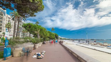 İnsanlar ünlü La Croisette Bulvarı yakınlarındaki limanda yürüyorlar. Fransız Rivierası. Yeşil ağaçlar, yaz gününde mavi bulutlu gökyüzü