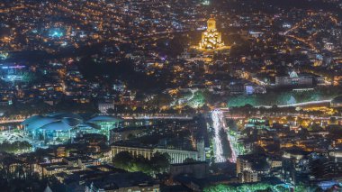 Aydınlanmış Tsminda Sameba Katedrali ya da Tiflis 'in Kutsal Üçlü Katedrali ve Kamu Hizmetleri Salonu hava gününden gece geçiş zamanına. Tiflis, Gürcistan 'daki ana katedral. Arka plandaki evler