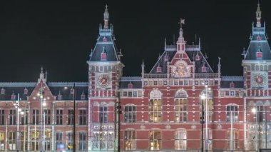 Aydınlatılmış Central Railway İstasyonu gece zaman çizelgesi. Hollanda, Amsterdam 'daki tren istasyonunun önündeki bir iskeleye, tramvaylara ve kanallara demirlemiş tur tekneleri