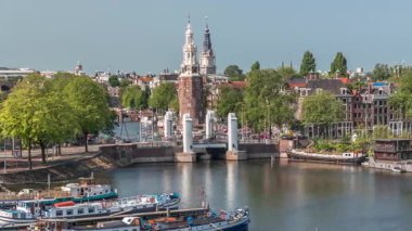 Amsterdam 'ın şehir panoramasının önünde Montelbaanstoren kulesi ve kanal boyunca uzanan şehir manzarası yer almaktadır. Eski kentin rıhtımında tarihi Hollanda evleri, mavnalar ve yüzen evler