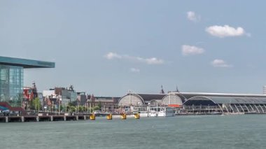 Amsterdam Merkez İstasyonu 'nun kıvrımlı çatısı ve Amstel Nehri zaman dilimi ile panoramik manzarası. Bir feribot, bulutların hareketli olduğu güneşli bir gökyüzünün altında nehri geçer. Amsterdam, Hollanda