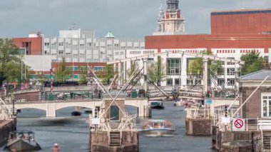 Amstel Nehri 'ndeki Amstel kilitlerinden geçen teknelerin zamanına göre görüntüsü. Opera binası ve Zuiderkerk kulesi arka planda görünüyor. Bulutlar gökyüzünü dolduruyor. Amsterdam, Hollanda