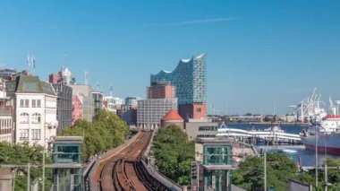 Almanya 'nın Hamburg kentindeki Landungsbrucken istasyonuna gelen trenler son sürat ilerliyor. Metroya binen insanlar mavi gökyüzünün altında Elbphilharmonie, liman ve marina ile panoramik manzara