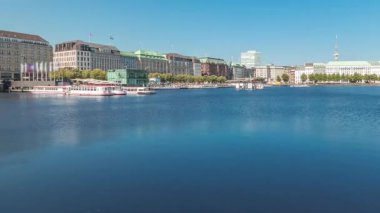 Panorama, Almanya, Hamburg 'da Binnenalster zaman çizelgesini tekne istasyonuyla gösteriyor. Alster Gölü 'nün iç sularına yansıyan tarihi binalar mavi gökyüzünün altında. İskelede kuğularla manzaralı şehir manzarası