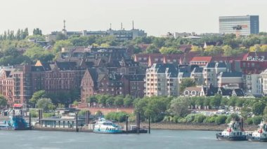Almanya 'nın Hamburg kentinde Elbe nehir kenarının hava zaman çizelgesi. İskeleye yakın tekneler, marina ve geçen gemilerle manzara. İnşaat alanında vinçleri olan evler. Elbe Nehri kıyısındaki canlı şehir manzarası