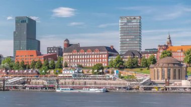 Hamburg limanına giriş ve çıkış yapan bir feribotun zaman çizelgesi. İskelede yolcuları var ve merdivenlerde ilerliyor. Arkaplanda Elbe nehri boyunca tarihi ve modern binalar var. Almanya