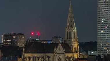Saint Servatius Kilisesi 'nin Belçika, Brüksel' deki Schaerbeek şehrindeki uçuş zamanı. Aydınlanmış evlerle çevrili Neo-Gotik kule. Arka planda yüksek binalar ve vinçler var