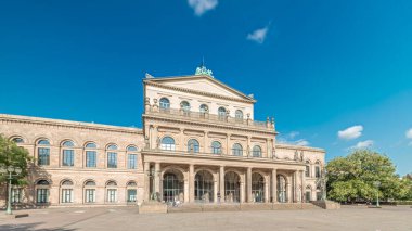 Hanover Devlet Operası 'nın (Staatsoper Hannover) ön görüntüsü. Şehir meydanında canlı mavi bulutlu bir zaman dilimi altında yürüyen insanların olduğu klasik tarzda bir tiyatro. Almanya