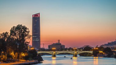 Guadalquivir Nehri yakınlarındaki Torre Sevilla gökdeleninin yer aldığı Seville 'in ufuk çizgisi gece gündüz geçiş zamanı. Aydınlatılmış Triana Köprüsü su, modern ve geleneksel mimariyi yansıtıyor
