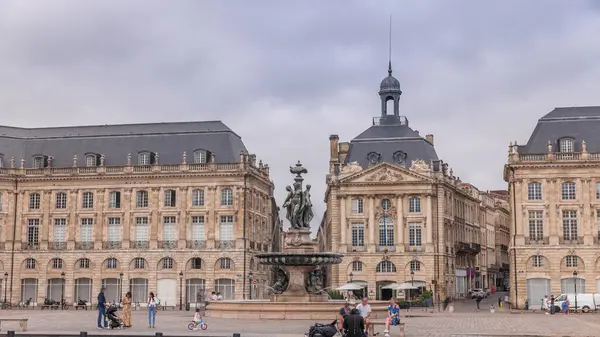 Place de la Bourse zaman atlaması Fransa 'nın Bordeaux şehrinde, Bulutlu mavi gökyüzünün altında klasik mimari ile çevrili Üç Zarafet Çeşmesi ile birlikte. İnsanlar ana meydanda bir tramvay durağında