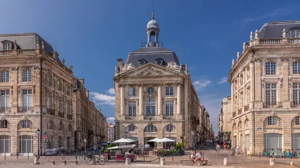 Place de la Bourse, Fransa 'nın Bordeaux şehrinde, bulutlu mavi gökyüzü altında klasik mimari ile çevrili restoranları olan bir yer. Meydanda ve ana caddelerde yürüyen insanlar