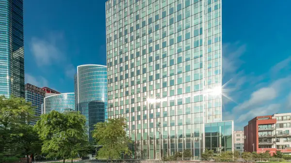 Panorama modern ofis binalarının zaman çizelgesini gösteriyor. Parktaki yeşil ağaçlar tarafından çerçevelenmiş parlak mavi gökyüzü üzerinde yansımaları ve röfleleri olan cam gökdelenler. Brüksel 'de güneş ışığı ve geçen bulutlar