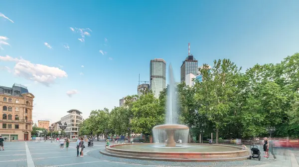 Frankfurt, Almanya merkezindeki Opera Meydanı (Opera Meydanı) zaman atlaması, arka planda gökdelenler ve kulelerle çevrili ve akşam gökyüzü altında ağaçlarla çevrili bir çeşme ve şehir hayatı sergiliyor.