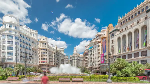 Valencia, İspanya 'da Belediye Meydanı' ndaki (Plaza del Ayuntamiento Meydanı) zaman atlaması. Büyük mimari, çiçek açan yeşillik ve bulutlu bir gökyüzünün altında su jetleri. İnsanlar canlı meydanda geziniyor..