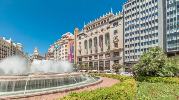 Panorama, İspanya 'nın Valencia şehrinde Belediye Meydanı' nda (Plaza del Ayuntamiento Meydanı) fıskiyeyi gösteriyor. Büyük mimari, çiçek açan yeşillik ve mavi gökyüzünün altında su jetleri. İnsanlar meydanda geziniyor.