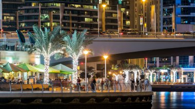 Gece vakti Dubai Marina 'da kafeler ve restoranlarla gezinti, BAE. Palmiyeler, tekneler ve kulelerle rıhtımın manzarası. Yapay kanallı bölge