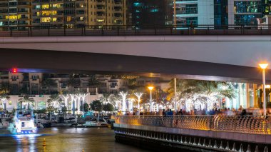 Yürüyen insanlarla dolaş ve Dubai Marina Kanalı üzerinde trafiği olan köprü gece vakti, BAE. Palmiyeler, tekneler ve kulelerle rıhtımın manzarası. Yapay kanallı bölge