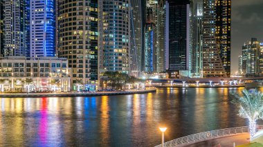 Dubai Marina 'da, Dubai gecesi, Birleşik Arap Emirlikleri' nin rıhtımından suya yansıyan tekneler, palmiyeler ve modern kuleler yer almaktadır. Gökdelende yapay kanalı ve aydınlatılmış pencereleri olan bir bölge