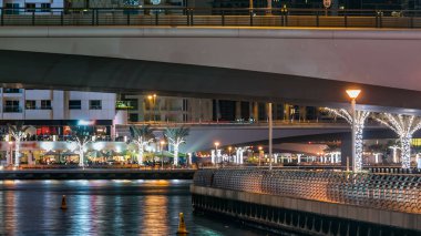 Gece vakti Dubai Marina 'da kafeler ve restoranlarla gezinti, BAE. Palmiyeler, tekneler ve kulelerle rıhtımın manzarası. Yapay kanallı bölge
