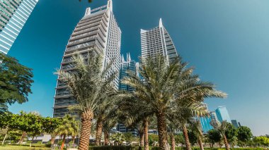 Parktaki gökdelenlerin palmiyeli görüntüsü. Dubai, BAE 'deki Jumeirah Gölü Kuleleri' nde camla kaplı konut binaları. Çimlerden görünüm