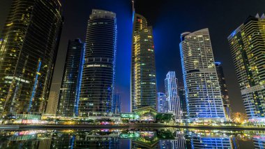 Rıhtımdaki ışıklandırılmış gökdelenlerin gece görüş açısını hızlandırın. Dubai, BAE 'deki Jumeirah Gölü Kuleleri' ndeki konut binaları. Rıhtımdan suya yansıyan görüntüyü göster