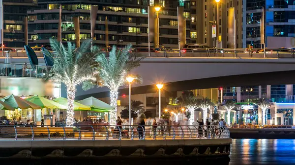 Gece vakti Dubai Marina 'da kafeler ve restoranlarla gezinti, BAE. Palmiyeler, tekneler ve kulelerle rıhtımın manzarası. Yapay kanallı bölge