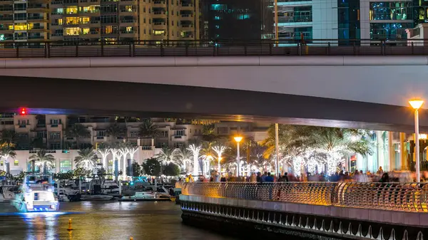 Yürüyen insanlarla dolaş ve Dubai Marina Kanalı üzerinde trafiği olan köprü gece vakti, BAE. Palmiyeler, tekneler ve kulelerle rıhtımın manzarası. Yapay kanallı bölge