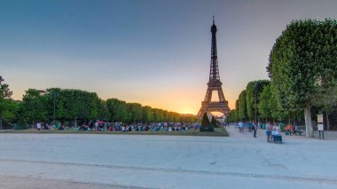 Güneş batarken Champ de Mars 'tan görülen Eyfel Kulesi, Paris, Fransa. İnsanlar çimenlerde oturup yürüyorlar. Açık gökyüzü ve güneşli bir yaz akşamı